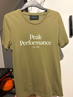 Olivgrön Peak Performance t-shirt - Snygg olivgrön t-shirt från Peak Performance med vit logga och text 'Est. 1986' på bröstet. Klassisk rund halsringning och korta ärmar. Perfekt för en avslappnad och stilren look.