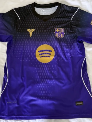 Lila FC Barcelona matchtröja - Snygg lila FC Barcelona matchtröja med guldfärgade detaljer och logotyper. Tröjan har svart överdel som tonar ner i lila, vita linjer längs sidorna och är tillverkad i ett lätt, ventilerande funktionsmaterial. Perfekt för fotbollsträning eller att bära som supporter.