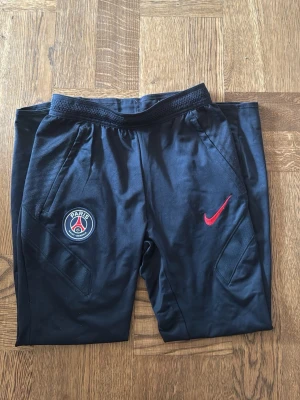 Svarta PSG träningsbyxor Nike stl: 137-147 - Svarta träningsbyxor från Nike med Paris Saint-Germain-logga på ena benet och röd Nike Swoosh på andra. Byxorna har smal passform, elastisk midja och dragkedjefickor. Perfekta för fotbollsträning eller chill. Materialet är troligtvis polyester.
