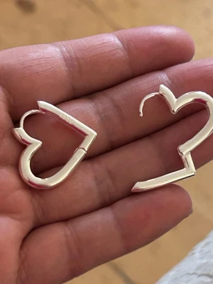 Silvriga hjärtformade örhängen - Säljer ett par stilrena örhängen i form av hjärtan. De är i blankt silver. metallmaterial och har en öppen design som ger en modern touch. Perfekta för dig som vill ha något unikt och lekfullt i smyckeskrinet.