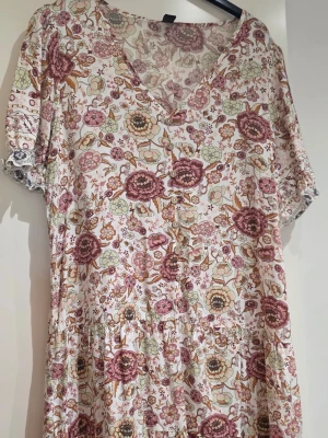 Blommig kortklänning med v-ringning - Söt kortklänning med v-ringning och korta ärmar. Klänningen har ett blommigt mönster i rosa, beige och grönt på vit botten samt knappar framtill. Tillverkad i mjuk bomull, perfekt för vår och sommar.
