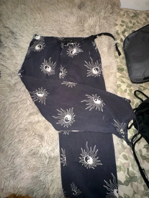 Svarta mjukisbyxor med yin yang-print - Svarta mjukisbyxor från Sweet SKTBS med vita sol- och yin yang-motiv över hela byxan. Byxorna har resår och dragsko i midjan för skön passform. Perfekta för en chill och avslappnad stil.