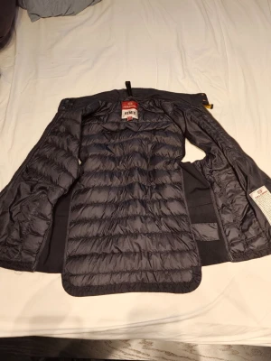 Svart dunjacka från Parajumpers - Snygg svart dunjacka från Parajumpers med quiltad insida och klassisk design. Jackan har en lätt glansig yta och detaljer som dragkedja och innerficka. Perfekt för kalla vinterdagar och riktigt varm tack vare dunfyllningen.
