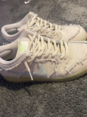 SB Low sneakers i beige canvas - Unika  SB Low sneakers i beige canvas med fransade detaljer och ljusgrå swoosh. Skorna har snörning och en ljus sula. Märkeslogga på plösen och färgglad innersula. Perfekta för dig som gillar streetwear och en avslappnad stil. Dem lyser också i mörkret.