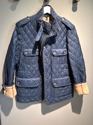 Blå quiltad jacka från Burberry Brit - Klassisk blå quiltad jacka från burberry brit med de ikoniska rutiga fodret. Jacka med dragkedja o knappar fram. Perfekt för dig som gillar tidlös o stilren design.