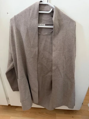 Beige kofta från Oysho - Mysig beige kofta från Oysho med öppen front och stora fickor. Koftan är stickad i ett mjukt material och har en avslappnad passform, perfekt att slänga på sig över en t-shirt eller topp. Enkel och stilren design som funkar året runt.