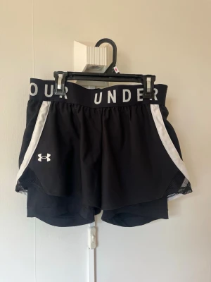 Svarta träningsshorts Under Armour play up 2-in-1 - Svarta träningsshorts från Under Armour med bred resår i midjan där loggan står tryckt runtom. Shortsen har vita detaljer längs sidorna och meshpaneler för extra ventilation. Perfekta för löpning eller gympass. Ficka på båda sidorna samt innerficka 
