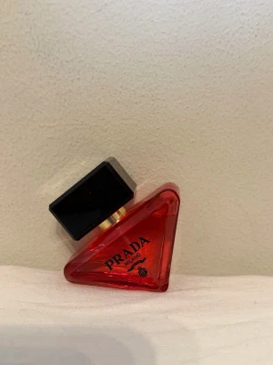 Prada parfym röd trekantig flaska - Snygg parfym från Prada i en unik trekantig flaska av rött glas med svart fyrkantigt lock och gulddetalj. Flaskan har en modern design och känns lyxig, perfekt för dig som gillar exklusiva dofter och vill ha något som sticker ut i badrumshyllan.