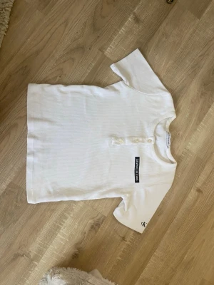 Vit ribbad t-shirt Calvin Klein Jeans - Vit ribbad t-shirt från Calvin Klein Jeans med korta ärmar och knappdetaljer vid halsen. T-shirten har en svart logotyp på bröstet och en liten CK-brodyr på ena ärmen. Tillverkad i mjuk bomull för en skön känsla.