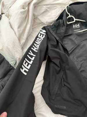 Svart Helly Hansen vindjacka - Svart vindjacka från Helly Hansen med vit logga på ärmen och meshfoder. Jackan har Helly Tech-material som är vattenavvisande och andas. Praktiska dragkedjefickor och luva. Perfekt för utomhusaktiviteter och sport.