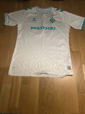 Vit Werder Bremen matchtröja - Snygg vit matchtröja från Werder Bremen med turkosa detaljer och nummer 14 på ryggen. Tröjan har klubbmärke på bröstet, sponsorlogga och diskret mönster i tyget. Perfekt för fotbollsträning eller att bära på läktaren.