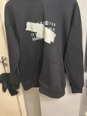 Svart Maison Margiela hoodie - Cool svart hoodie från Maison Margiela med vit grafisk print och tejpdetalj framtill. Klassisk känguruficka och huva med dragsko. Perfekt för dig som gillar streetwear och unika designer.