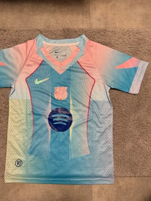FC Barcelona x Nike pastelltröja - Säljer en FC Barcelona matchtröja från Nike i pastellfärger med blå, rosa och gula toner. Tröjan har klubbmärke, Nike-logga och Spotify-logga på bröstet. Tillverkad i lätt och ventilerande polyester, perfekt för fotboll eller träning.