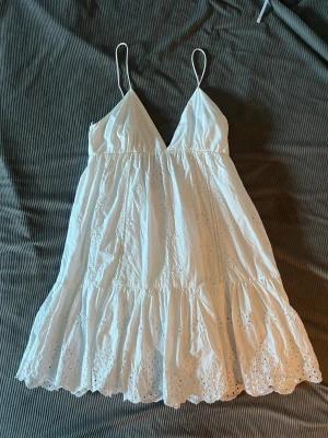 Vit sommarklänning med broderi  - Söt vit kortklänning med tunna axelband och v-ringning. Klänningen har volangkant nertill och fina broderade detaljer i tyget. Perfekt för varma dagar och har en luftig passform. Storleken är M men skulle nog sagt S. Skriv för fler bilder eller andra funderingar! 