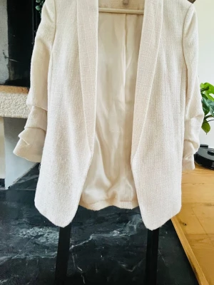 Vit kavaj från H&M, storlek 38 - Stilren vit kavaj från H&M i storlek 38. Kavajen har en rak passform, uppvikta ärmar och är tillverkad i ett strukturerat tyg med fodrad insida. Perfekt för en trendig och fräsch look.