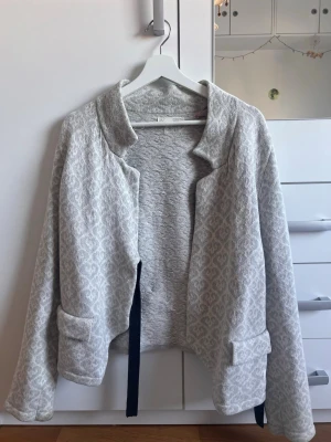 Ljusgrå mönstrad cardigan Odd Molly - Snygg ljusgrå cardiganjacka från Odd Molly med diskret vitt mönster. Jackan har öppen front, två fickor och ett svart knytband. Mjuk stickad kvalitet och broderad Odd Molly-logga på ärmen ger en unik touch.