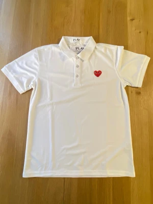 Vit Comme des Garçons PLAY t-shirt - Vit t-shirt från Comme des Garçons PLAY med klassisk pikékrage och knappar framtill. På bröstet sitter det ikoniska röda hjärtat med ögon. T-shirten är kortärmad och har en clean, stilren look. Tillverkad i mjuk bomull för skön känsla.