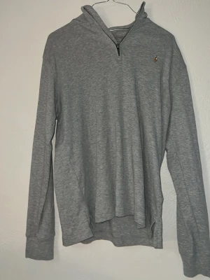 Grå hoodie från Polo Ralph Lauren - Snygg grå hoodie från Polo Ralph Lauren med dragkedja vid halsen och liten broderad logga på bröstet. Tillverkad i mjukt material och har långa ärmar samt huva. Perfekt för en avslappnad och stilren look.