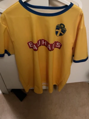 Svensk landslagströja gul nr 22 - Gul svensk landslagströja med blå detaljer, Bauhaus-logga på bröstet och ryggen samt nummer 22 på ryggen. Broderad svensk landslagssköld på bröstet. Perfekt för dig som vill visa ditt stöd för Sverige på matchen eller träningen.