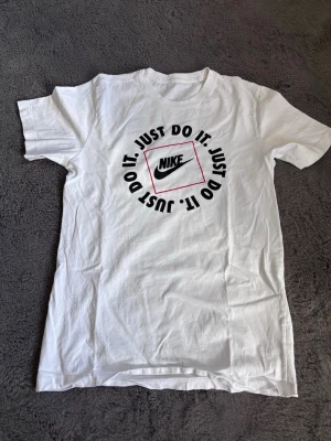 Nike - Vit t-shirt från Nike med det klassiska 'Just Do It.'-trycket i svart och rött på bröstet. T-shirten har rund halsringning och korta ärmar. Perfekt för en sportig och avslappnad stil.