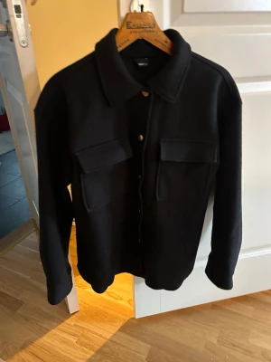 Gina Tricot skjorta/ jacka i storlek xs - Stilren svart overshirt med klassisk krage och tryckknappar framtill. Två stora bröstfickor med lock ger en cool och praktisk look. Overshirten har raka ärmar och är tillverkad i ett tjockare tyg som passar perfekt till lager-på-lager-stil..