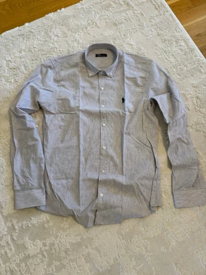 Ljusgrå skjorta från Ralph Lauren - Klassisk ljusgrå skjorta från Ralph Lauren med button-down krage och svart broderad logga på bröstet. Skjortan har långa ärmar och vita knappar framtill. Perfekt för en stilren look.