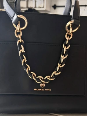 Svart handväska från Michael Kors - Stilren svart handväska från Michael Kors med guldfärgade detaljer och kedjehandtag. Väskan har dragkedja upptill och är tillverkad i slätt läder. Perfekt för dig som gillar klassisk och lyxig design.köperan står för frakten.
