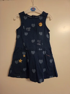 Blå jeansklänning med hjärtan, stl 4-5 år 110 cm - Söt blå jeansklänning med ljusa hjärtmönster, ärmlös design och rund hals. Klänningen har broderade detaljer som stjärnor och text, samt en liten applikation framtill. Perfekt för en lekfull och trendig look.