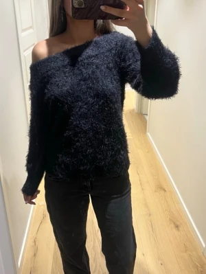 Svart fluffig off shoulder stickad tröja - Mysig svart stickad tröja med fluffig textur och off shoulder-design. Tröjan har långa ärmar och en avslappnad passform som ger en trendig look. Perfekt för dig som gillar att sticka ut med en unik och bekväm stil.