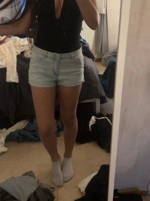 Ljusblå denimshorts med normal passform - Säljer ett par ljusblå denimshorts med klassisk femficksdesign och normal passform. Shortsen har knapp och dragkedja framtill samt bälteshällor. Perfekta för varma dagar och enkla att matcha med olika toppar.