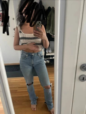 Ljusblå ripped straight leg jeans - Säljer ett par ljusblå jeans med raka ben och slitningar vid knäna. Jeansen har hög midja, klassiska fem fickor och knappgylf. Perfekta för en avslappnad och trendig look. Materialet är denim i bomull.