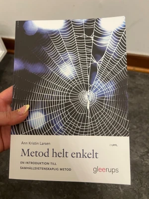 Metod helt enkelt kursbok - Kurslitteratur: 'Metod helt enkelt' av Ann Kristin Larsen, 2:a upplagan. Boken har ett omslag med ett spindelnät i blått och vitt och är tryckt på matt papper. Perfekt för dig som pluggar samhällsvetenskapliga metoder.
