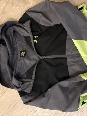 Under Armour träningsjacka grå/svart - Snygg träningsjacka från Under Armour i grått, svart och neongult. Jackan har hel dragkedja, huva och meshfoder för extra ventilation. Perfekt för löpning eller utomhusträning. Under Armour-logga i neongult på bröstet.