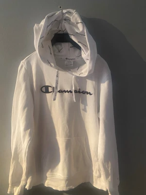 Vit hoodie från Champion med logga, ungdom - Säljer en vit hoodie från Champion med stor mörkblå logga på bröstet och tryckt logomönster i huvan. Klassisk känguruficka framtill och dragsko i huvan. Strl L, ungdom 