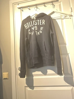 Svart hoodie från Hollister - Svart hoodie från Hollister med vit text 'Hollister 49 Surf Team' på bröstet. Klassisk känguruficka framtill och vit dragsko i huvan. Perfekt för en avslappnad och sportig stil.