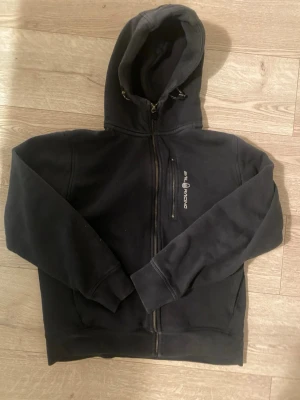 Svart zip-hoodie från Sea Racing - Svart hoodie med dragkedja från Sea Racing. Har en huva med dragsko och en bröstficka med dragkedja. Mjuk insida och ribbade muddar vid ärmslut och nederkant. Perfekt för en avslappnad stil.