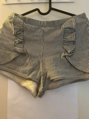 Randiga shorts med volangdetaljer - Snygga svartvita randiga shorts med volangdetaljer på sidorna och elastisk midja. Perfekta för varma dagar och ger en trendig look. Tillverkade i mjukt bomullstyg som känns skönt mot huden.