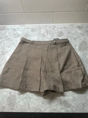 Rutiga skorts i brunbeige ton - Snygga skorts i brunbeige rutigt mönster med två dekorativa knappar framtill. Klassisk A-linjeform och diskret plissering framtill. Perfekt för en trendig och stilren look. Materialet ser ut att vara en mjuk vävd textil.