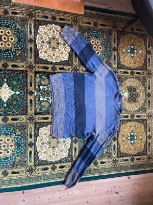 Blå- och grårandig stickad tröja XL - Stickad tröja från Chaps Ralph Lauren i storlek XL. Tröjan har breda horisontella ränder i blått och grått, rund halsringning och ribbstickade muddar. Tillverkad i 100% bomull, perfekt för lager-på-lager-stil.