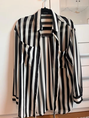 Randig skjorta från Bershka - Snygg skjorta från Bershka med svarta och vita vertikala ränder. Skjortan har lång ärm, klassisk krage och två bröstfickor. Tillverkad i ett lätt och mjukt polyestertyg som ger en skön känsla och cool look.