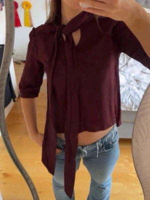Vinröd blus med knytband i halsen - Trendig vinröd blus med knytband i halsen och snygg v-ringning. Blusen har trekvartsärm och är tillverkad i ett lätt, flowy tyg som ger en avslappnad men stilren look. Perfekt att matcha med jeans eller kjol för en cool outfit.