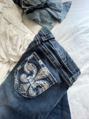 Mörkblå jeans med broderad ficka - Säljer ett par mörkblå jeans med en stor, broderad ficka bak i beige och blått mönster. Lånade bilder! Säljer då de inte kommer till användning, knappt använda