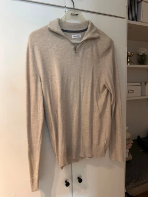 Beige tunn stickad tröja - En stilren och tunnstickad beige tröja med långa ärmar och ribbade muddar. Perfekt för lager-på-lager och enkel att matcha med olika outfits. Tröjan har en klassisk passform och mjukt material som känns skönt mot huden.