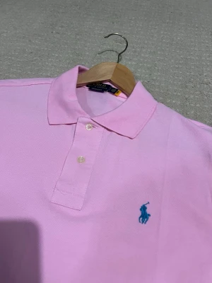Rosa pikétröja, Ralph Lauren - Snygg ljusrosa pikétröja från Ralph Lauren med klassisk krage och två knappar framtill. Tröjan har korta ärmar och den ikoniska blå loggan broderad på bröstet. Perfekt för en stilren och avslappnad look.