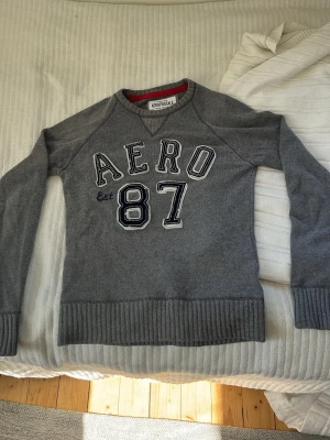 Grå tröja från Aeropostale - Grå sweatshirt från Aeropostale med broderad text 'AERO 87' på bröstet. Tröjan har rund hals, långa ärmar och ribbade muddar vid ärmslut och nederkant. Materialet är 60% bomull och 40% akryl. Perfekt för en avslappnad och sportig stil.