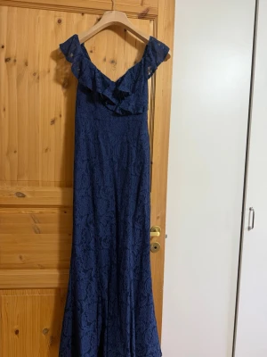 Marinblå långklänning med volang - Elegant marinblå långklänning med offshoulder och volangdetaljer upptill. Klänningen har ett vackert spetsmönster och är figurnära upptill med utsvängd nederdel. Perfekt för speciella tillfällen när du vill känna dig extra fin.