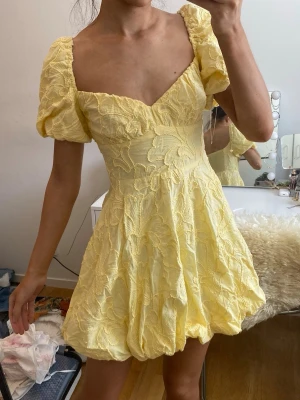 Gul kort klänning med puffärmar - Superfin gul kort klänning med puffärmar och v-ringning. Klänningen har broderade blommor och en voluminös kjol med ballongformad nederkant. Perfekt för sommarens alla tillfällen! Tyget runt dragkedjan är lite trasigt men den funkar perfekt fortfarande. Köpt för 600