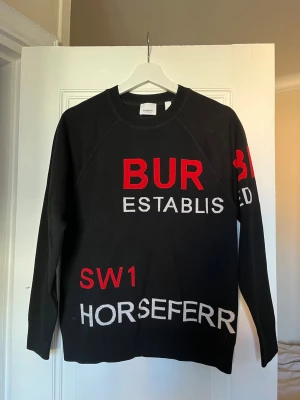 Black Burberry sweatshirt horseferry print - Svart sweatshirt från Burberry med stora röda och vita bokstäver och textdetaljer på både fram- och baksida. Rund halsringning och ribbade muddar. Använd 2 gånger. Nypris ca 6800kr.