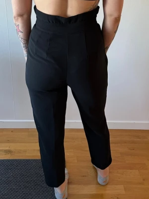 Svarta high waist kostymbyxor - Snygga svarta kostymbyxor med hög midja och raka ben. Byxorna har två dekorativa knappar i midjan och pressveck framtill. Modellen är croppad och har en modern, stilren look. Perfekta för dig som gillar trendiga och bekväma byxor.