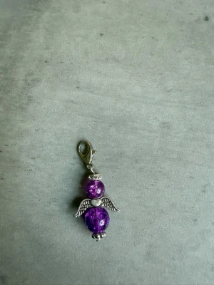 Lila ängelberlock med vingar - Söt berlock formad som en ängel med två lila glaspärlor, silverfärgade detaljer och små vingar. Perfekt att fästa på armband, halsband eller väska. Karbinhake i metall för enkel fastsättning. Handgjord av mig!